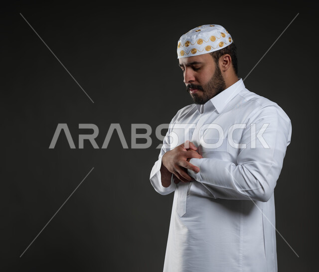 أداء فرائض الصلاة بأوقاتها، مفهوم أركان الإسلام، بورتريه مقرب لرجل عربي خليجي سعودي مسلم يرتدي الثوب والقبعة الإسلامية البيضاء يقف على السجادة، العبادة والتقرب إلى الله، الركوع و السجود والخشوع، قراءة ما تيسر من القرآن الكريم، خلفية رمادية