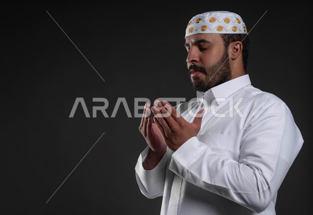 أداء فرائض الصلاة بأوقاتها، شاب وسيم مسلم، الدعاء و التضرع لرب العباد، مفهوم أركان الإسلام، بورتريه مقرب لرجل عربي خليجي سعودي يرتدي الثوب والقبعة الإسلامية البيضاء يقف على السجادة، العبادة والتقرب إلى الله، خلفية رمادية