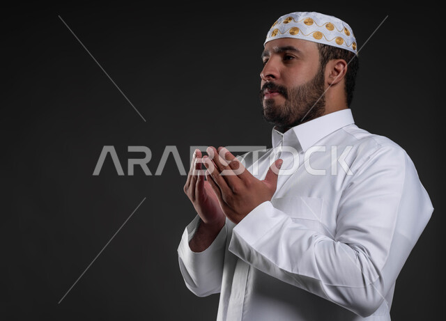 الدعاء و التضرع لرب العباد، أداء فرائض الصلاة بأوقاتها، مفهوم أركان الإسلام، بورتريه مقرب لرجل عربي خليجي سعودي يرتدي الثوب والقبعة الإسلامية البيضاء يقف على السجادة، شاب وسيم مسلم، العبادة والتقرب إلى الله، خلفية رمادية