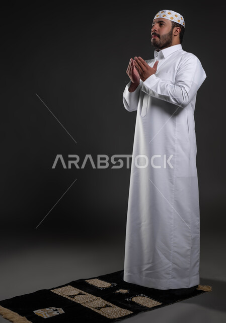 Worship and closeness to God, performing the obligatory prayers on time, a handsome young Muslim man, supplication and supplication to the Lord of servants, the concept of the pillars of Islam, a portrait of a Saudi Gulf Arab man wearing the white Islamic thobe and hat standing on the carpet, full-length body portrait, gray background