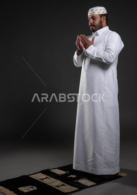 Supplication and supplication to the Lord of servants, performing the obligatory prayers at their appointed times, the concept of the pillars of Islam, a portrait of a Saudi Gulf Arab man wearing the white Islamic dress and hat standing on the carpet, a handsome young Muslim man, worship and getting closer to God, a full-body portrait, gray background