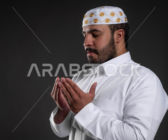 شاب وسيم مسلم، الدعاء و التضرع لرب العباد، أداء فرائض الصلاة بأوقاتها، مفهوم أركان الإسلام، بورتريه مقرب لرجل عربي خليجي سعودي يرتدي الثوب والقبعة الإسلامية البيضاء يقف على السجادة، العبادة والتقرب إلى الله، خلفية رمادية
