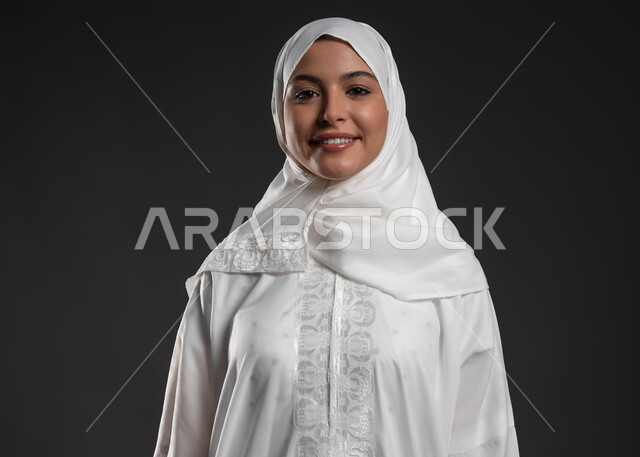 Looking at the camera with gestures of happiness and pleasure, the concept of commitment to religion, a close-up portrait of a Saudi Gulf Arab woman wearing the hijab and white abaya, standing upright, expressions of contentment and reassurance, a beautiful young Muslim woman, worship and closeness to God Almighty, gray background