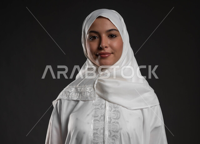 Looking at the camera with gestures of reassurance and contentment, the concept of adherence to religion, a close-up portrait of a Saudi Gulf Arab woman wearing the hijab and white abaya, standing upright, expressions of happiness and pleasure, a beautiful young Muslim woman, worship and getting closer to God Almighty, gray background
