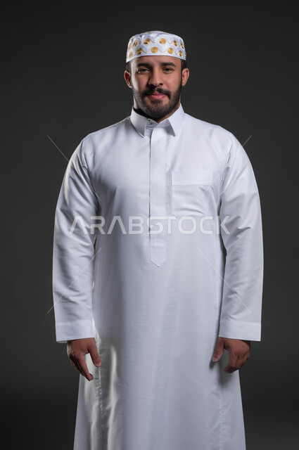 شاب مسلم وسيم، مفهوم الالتزم بالدين، بورتريه لرجل عربي خليجي سعودي يرتدي الثوب و القبعة الإسلامية البيضاء يقف باستقامة ينظر إلى الكاميرا بإيماءات الرضا و الطمأنينة، العبادة و التقرب إلى الله تعالى، خلفية رمادية