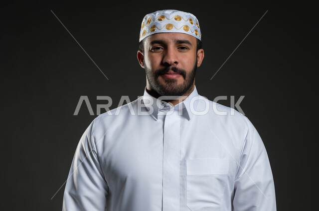 مفهوم الالتزم بالدين، بورتريه مقرب لرجل عربي خليجي سعودي يرتدي الثوب و القبعة الإسلامية البيضاء يقف باستقامة ينظر إلى الكاميرا بإيماءات الرضا و الطمأنينة، شاب وسيم مسلم، العبادة و التقرب إلى الله تعالى، خلفية رمادية