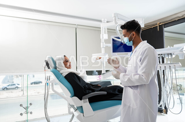 تقديم الخدمات الصحية، مفهوم الطب والرعاية الصحية,  الاهتمام في الصحة الفموية, طبيب أسنان عربي خليجي سعودي يرتدي المعطف و القفازات والكمامة الطبية يعطي نصائح للمحافظة على صحة الفم والأسنان لمريضة عربية خليجية سعودية محجبة, ايماءات اليدين