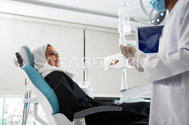 النظر الى الطبيب بايماءات السرور والسعادة, الارشادات والتعليمات الطبية, الاهتمام في الصحة الفموية, طبيب أسنان عربي خليجي سعودي يرتدي المعطف و القفازات والكمامة الطبية يعطي نصائح طبية لامراة عربية خليجية سعودية ترتدي الزي التقليدي تجلس في العيادة السنية