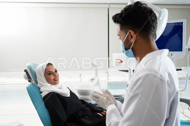 الإرشادات والنصائح الطبية, تقديم الخدمات الصحية، مفهوم الطب والرعاية الصحية,  الاهتمام في الصحة الفموية, طبيب أسنان عربي خليجي سعودي يرتدي المعطف و القفازات والكمامة الطبية يعطي نصائح للمحافظة على صحة الفم والأسنان لمريضة عربية خليجية سعودية محجبة