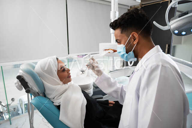 الأساليب العلاجية المتبعة للعناية بالفم, استخدام الأدوات الطبية السنية المعقمة، طبيب أسنان عربي خليجي سعودي يرتدي المعطف والقفازات والكمامة الطبية يمسك بيده ادوات طبية، سيدة عربية خليجية سعودية في عيادة سنية تتلقى العلاج اللازم