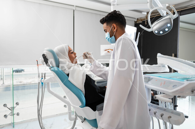 مفهوم الصحة الفموية, معالجة التسوس وتركيب الحشوات، جراحة الفم والأسنان، طبيب أسنان عربي خليجي سعودي يقوم بفحص أسنان المريضة و تشخيص المرض، سيدة عربية خليجية سعودية محجبة ترتدي الزي التقليدي و تجلس في العيادة السنية تقوم بإجراء الفحوصات الدورية للأسنان