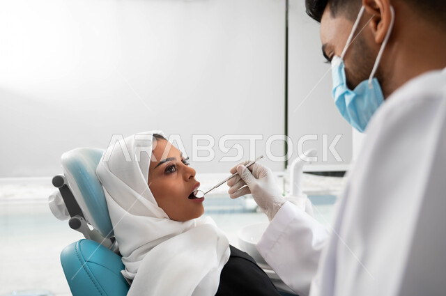 معالجة التسوس وتركيب الحشوات، جراحة الفم والأسنان، صورة مقربة لطبيب أسنان عربي خليجي سعودي يقوم بفحص أسنان المريضة و تشخيص المرض، سيدة عربية خليجية سعودية محجبة ترتدي الزي التقليدي و تجلس في العيادة السنية تقوم بإجراء الفحوصات الدورية للعناية بالصحة الفموية