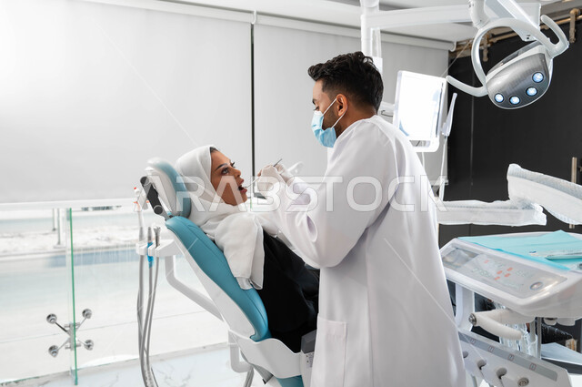 جراحة الفم والأسنان، صورة جانبية لطبيب أسنان عربي خليجي سعودي يقوم بفحص أسنان المريضة و تشخيص المرض، معالجة التسوس وتركيب الحشوات، سيدة عربية خليجية سعودية محجبة ترتدي الزي التقليدي و تجلس في العيادة السنية تقوم بإجراء الفحوصات الدورية للعناية بالصحة الفموية