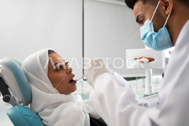Visiting health clinics and taking the necessary measures, a close-up picture of a Saudi Gulf Arab dentist wearing a coat, gloves and a medical mask holding a medical tool in his hand, a veiled Saudi Gulf Arab woman in a dental clinic, dental equipment and tools