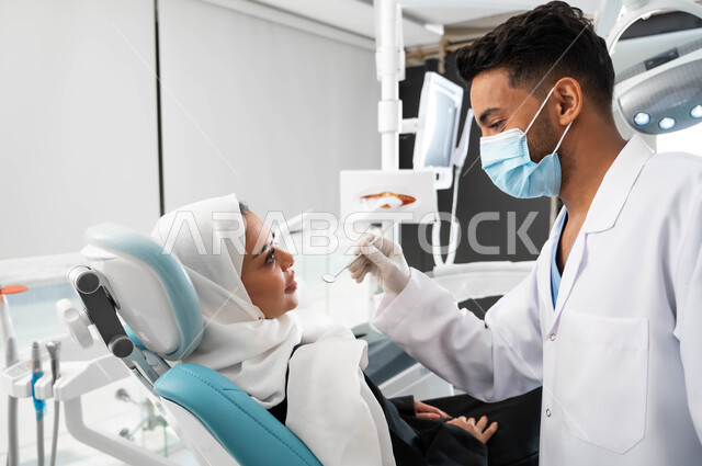 معدات وأدوات طب الأسنان، زيارة العيادات الصحية واتخاذ الإجراءات اللازمة، طبيب أسنان عربي خليجي سعودي يرتدي المعطف والقفازات والكمامة الطبية يمسك بيده أداة طبية، سيدة عربية خليجية سعودية محجبة في عيادة سنية