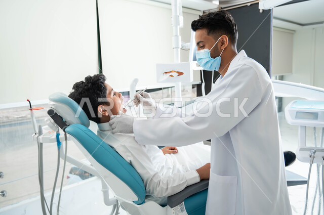 جراحة الفم والأسنان، صورة جانبية لطبيب أسنان عربي خليجي سعودي يقوم بفحص أسنان فتى و تشخيص المرض، معالجة التسوس وتركيب الحشوات، طفل عربي خليجي سعودي يرتدي الزي التقليدي و يجلس في العيادة السنية يقوم بإجراء الفحوصات الدورية للعناية بالصحة الفموية