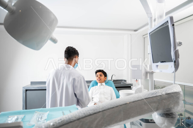 النظر الى الطبيب بايماءات السرور والسعادة, الارشادات والتعليمات الطبية, الاهتمام في الصحة الفموية, طبيب أسنان عربي خليجي سعودي يرتدي المعطف و القفازات والكمامة الطبية يعطي نصائح طبية لطفل عربي خليجي سعودي يرتدي الزي التقليدي يجلس في العيادة السنية