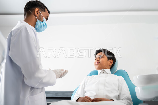 زيارة العيادات الصحية واتخاذ الإجراءات اللازمة, طبيب أسنان عربي خليجي سعودي يقوم بفحص أسنان المريض و تشخيص المرض، طفل عربي خليجي سعودي يرتدي الزي التقليدي و يجلس في العيادة السنية يقوم بإجراء الفحوصات الدورية للعناية بالصحة الفموية