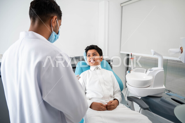 الارشادات والتعليمات الطبية, الاهتمام في الصحة الفموية, النظر الى الطبيب بايماءات السرور, طبيب أسنان عربي خليجي سعودي يترتدي المعطف و القفازات والكمامة الطبية يعطي نصائح طبية لطفل عربي خليجي سعودي يرتدي الزي التقليدي يجلس في العيادة السنية