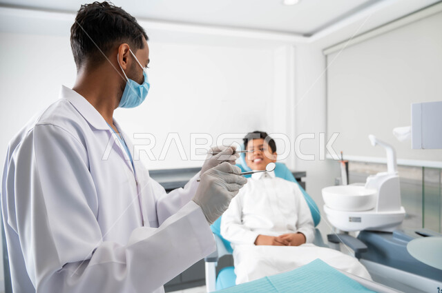 مفهوم العناية الدائمة بالأسنان, معالجة التسوس وتركيب الحشوات، طفل عربي خليجي سعودي في عيادة سنية لتقديم الخدممات الصحية, طب وجراحة الفم والأسنان، طبيب أسنان عربي خليجي سعودي يرتدي القفازات والكمامة الطبية يقوم بفحص وتشخيص أسنان طفل مريض