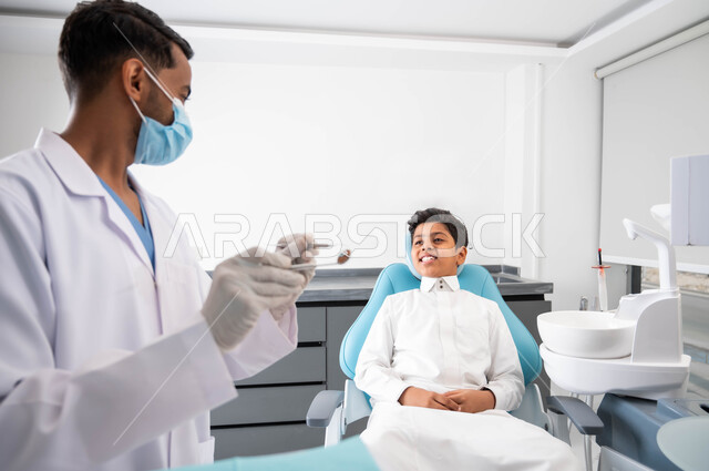 فتى سعودي يقوم بالزيارة الدورية للعيادة السنية للاهتمام بصحة الفم، الرعاية الصحية للأطفال، طبيب أسنان عربي خليجي سعودي يرتدي المعطف والقفازات والكمامة الطبية يقوم بفحص وتشخيص أسنان الولد