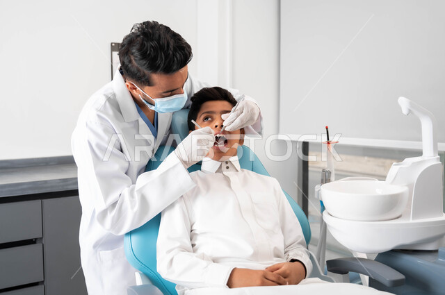 استخدام الأدوات الطبية السنية المعقمة، طبيب أسنان عربي خليجي سعودي يرتدي المعطف والقفازات والكمامة الطبية يمسك بيده ادوات طبية، فتى عربي خليجي سعودي في عيادة سنية يتلقى العلاج اللازم, الأساليب العلاجية المتبعة للعناية بالفم