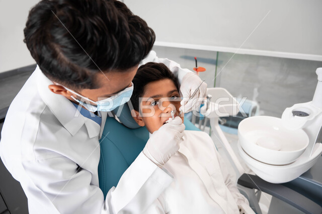 فتى سعودي يقوم بالزيارة الدورية للعيادة السنية للحفاظ على الأسنان من التسوس، مفهوم الطب والرعاية الصحية للأطفال، طبيب أسنان عربي خليجي سعودي يرتدي المعطف والقفازات والكمامة الطبية يقوم بفحص وتشخيص أسنان الولد