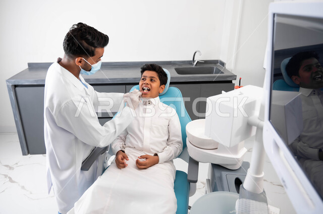 مفهوم الطب والرعاية الصحية للأطفال، فتى سعودي يقوم بالزيارة الدورية للعيادة السنية للحفاظ على الأسنان من التسوس، طبيب أسنان عربي خليجي سعودي يرتدي المعطف والقفازات والكمامة الطبية يقوم بفحص وتشخيص أسنان الولد