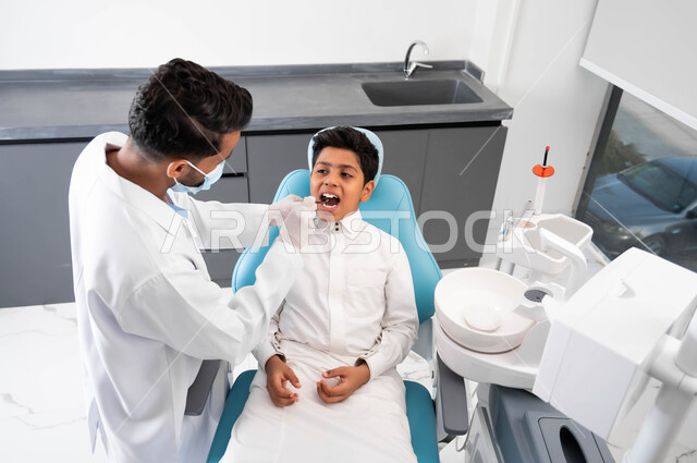 التأكد من سلامة الأسنان واللثة، المعدات والأدوات المعقمة، مفهوم المحافظة على الصحة الفموية، طبيب أسنان عربي خليجي سعودي يرتدي المعطف والقفازات والكمامة الطبية يقوم بفحص المريض، ولد يجلس في العيادة السنية يستعد لتلقي العلاج اللازم