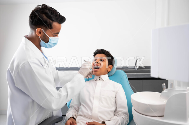 التأكد من سلامة الأسنان واللثة، المعدات والأدوات المعقمة، مفهوم المحافظة على الصحة الفموية، طبيب أسنان عربي خليجي سعودي يرتدي المعطف والقفازات والكمامة الطبية يقوم بفحص المريض، ولد يجلس في العيادة السنية يستعد لتلقي العلاج اللازم