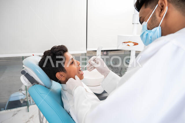 طبيب أسنان عربي خليجي سعودي يرتدي المعطف والقفازات والكمامة الطبية يقوم بفحص المريض، ولد يجلس في العيادة السنية يستعد لتلقي العلاج اللازم، معالجة الأسنان من النخور والتسوس، المعدات والأدوات المعقمة، المحافظة على الصحة الفموية