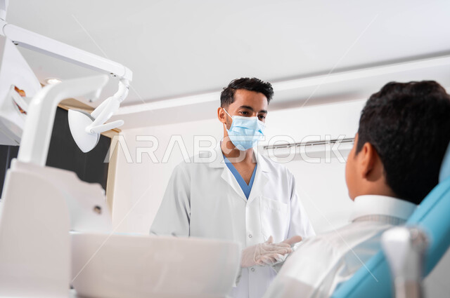 Medical instructions and instructions, a dentist talking to his patient with reassuring gestures, a Saudi Gulf Arab boy sitting in a dental clinic receiving advice, care and attention to oral health, treating teeth for caries and decay, periodic visits to health clinics