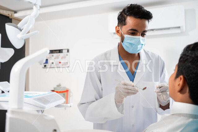 المعدات والأدوات المعقمة، مفهوم المحافظة على الصحة الفموية، زيارة العيادات الصحية لاتخاذ الإجراءات اللازمة، طبيب أسنان عربي خليجي سعودي يرتدي المعطف والقفازات والكمامة الطبية يمسك بيده آداة الفحص، ولد يجلس في العيادة السنية يستعد لتلقي العلاج اللازم