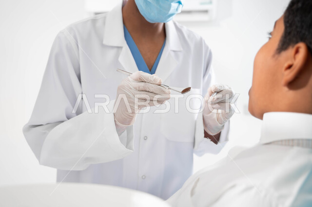 المعدات والأدوات المعقمة، مفهوم المحافظة على الصحة الفموية، زيارة العيادات الصحية لاتخاذ الإجراءات اللازمة، طبيب أسنان عربي خليجي سعودي يرتدي المعطف والقفازات والكمامة الطبية يمسك بيده آداة الفحص، ولد يجلس في العيادة السنية يستعد لتلقي العلاج اللازم