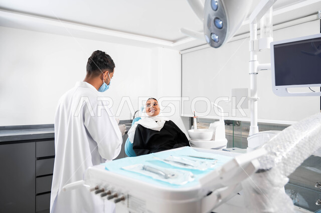 الإرشادات والتعليمات الطبية، أدوات ومعدات طب الأسنان المعقمة، طبيب أسنان عربي خليجي سعودي يتناقش مع مريضته، إمرأة عربية خليجية سعودية محجبة تجلس في عيادة سنية تنظر بإيماءات السرورتتلقى النصائح، العناية والاهتمام بالصحة الفموية