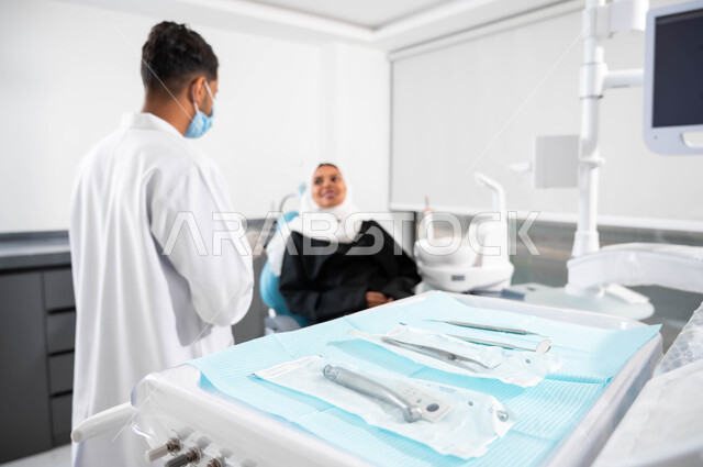الإرشادات والتعليمات الطبية، أدوات ومعدات طب الأسنان المعقمة، طبيب أسنان عربي خليجي سعودي يتناقش مع مريضته، إمرأة عربية خليجية سعودية محجبة تجلس في عيادة سنية تنظر بإيماءات السرورتتلقى النصائح، العناية والاهتمام بالصحة الفموية