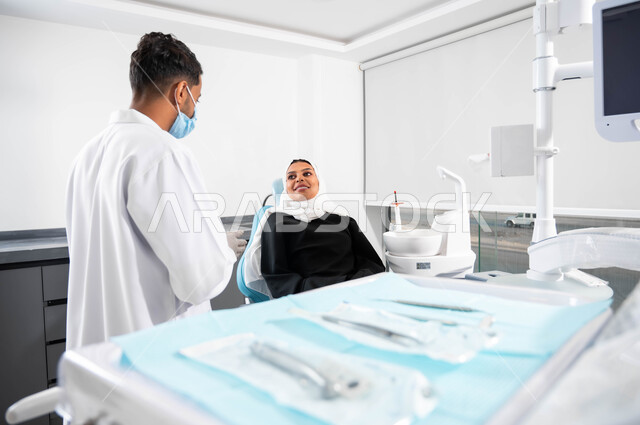 الإرشادات والتعليمات الطبية، أدوات ومعدات طب الأسنان المعقمة، طبيب أسنان عربي خليجي سعودي يتناقش مع مريضته، إمرأة عربية خليجية سعودية محجبة تجلس في عيادة سنية تنظر بإيماءات السرورتتلقى النصائح، العناية والاهتمام بالصحة الفموية