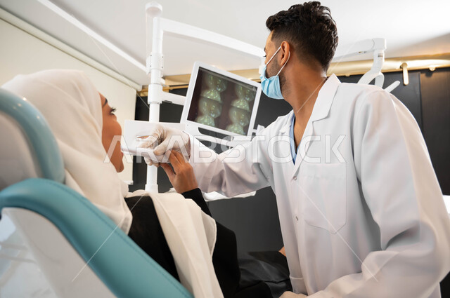 معدات وأدوات طب الأسنان، استخدام صور الأشعة السينية، طبيب أسنان عربي خليجي سعودي يرتدي المعطف والقفازات والكمامة الطبية يمسك بيده أداة يقوم بفحص مريضته،   سيدة محجبة تجلس في عيادة سنية تتلقى العلاج، العناية والاهتمام بصحة الفم، الزيارة الدورية للعيادات الصحية