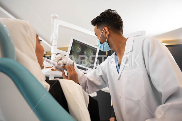 استخدام صور الأشعة السينية، معدات وأدوات طب الأسنان، طبيب أسنان عربي خليجي سعودي يرتدي المعطف والقفازات والكمامة الطبية يمسك بيده أداة الفحص،  سيدة محجبة تجلس في عيادة سنية تتلقى العلاج، العناية والاهتمام بصحة الفم، الزيارة الدورية للعيادات الصحية