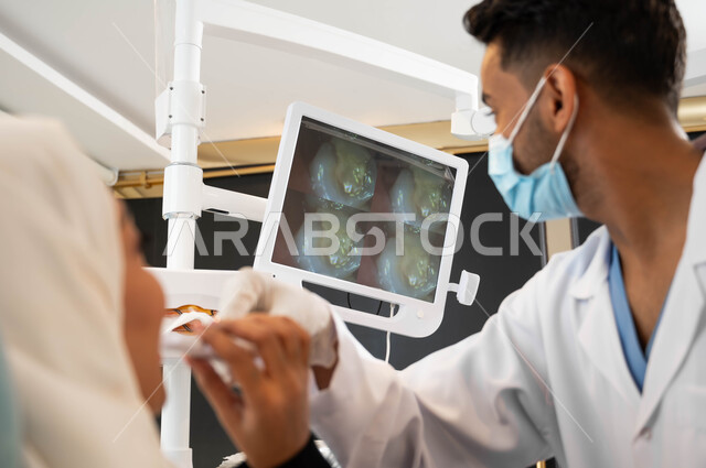 Using X-ray images, dental equipment and tools, a Saudi Arabian Gulf dentist wearing a coat, gloves and a medical mask holds an examination tool in his hand,   A veiled woman sits in a dental clinic receiving treatment, oral health care, and regular visits to health clinics