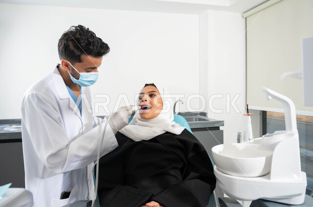 الزيارة الدورية للعيادات الصحية، معدات وأدوات طب الأسنان، صورة مقربة لطبيب أسنان عربي خليجي سعودي يرتدي المعطف والقفازات الطبية يمسك بيده أداة حفر السن وإزالة التسوس، سيدة عربية خليجية سعودية محجبة تجلس في عيادة سنية، العناية والاهتمام بصحة الفم
