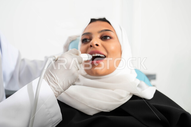 الزيارة الدورية للعيادات الصحية، معدات وأدوات طب الأسنان، صورة مقربة ليد طبيب أسنان يرتدي القفازات الطبية يمسك بيده أداة حفر السن وإزالة التسوس، سيدة عربية خليجية سعودية محجبة تجلس في عيادة سنية تتلقى العلاج، العناية والاهتمام بصحة الفم