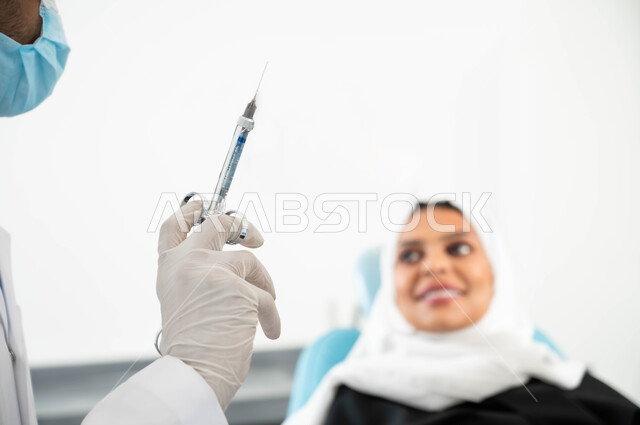 Local anesthetic injections to relieve pain, oral care using therapeutic methods, a close-up of a dentist’s hand wearing medical gloves holding an anesthesia needle, a veiled Saudi Arabian Gulf woman sitting in a dental clinic receiving the necessary treatmen