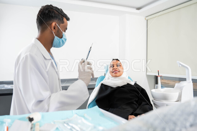Oral care using therapeutic methods, local anesthetic injections to relieve pain, a close-up picture of a Saudi Arabian Gulf dentist wearing a coat, gloves and a medical mask holding an anesthesia needle in his hand, a veiled woman in a dental clinic receiving the necessary treatment