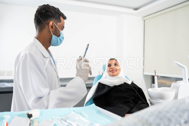 العناية بالفم بالطرائق العلاجية، حقن البنج الموضعي لتخفيف الإحساس بالألم، صورة مقربة لطبيب أسنان عربي خليجي سعودي يرتدي المعطف والقفازات والكمامة الطبية يمسك بيده إبرة التخدير، إمرأة محجبة في عيادة سنية تتلقى العلاج اللازم