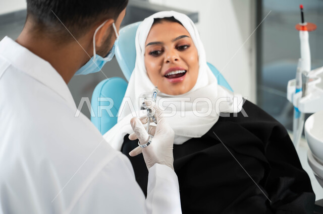 مفهوم الاهتمام والعناية بالصحة الفموية، طبيب أسنان يرتدي المعطف والقفازات والكمامة الطبية يستعد لحقن البنج الموضعي لتخفيف الألم، سيدة عربية خليجية سعودية محجبة تجلس في العيادة السنية تنظر الى الإبرة بإيماءات الخوف، تلقي العلاج واتخاذ الإجراءات اللازمة