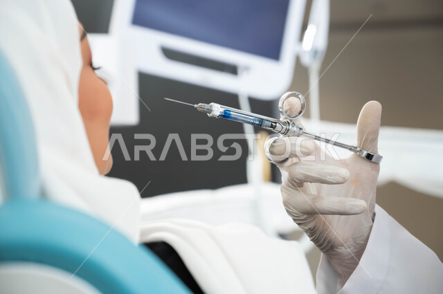 مفهوم العناية والاهتمام بالصحة الفموية، صورة مقربة ليد طبيب أسنان يرتدي القفازات الطبية يستعد لحقن البنج الموضعي لتخفيف الألم، سيدة محجبة تجلس في العيادة السنية تتلقى العلاج اللازم، زيارة العيادات الصحية واتخاذ الإجراءات اللازمة