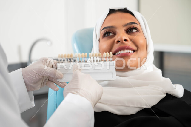 Using porcelain veneers, taking care of the health and appearance of the teeth, a close-up of a dentist wearing medical gloves holding a color guide to restore the patient’s teeth, a veiled Saudi Gulf Arab woman in a dental clinic receiving the necessary treatment to create a distinctive smile