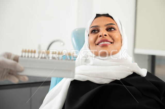 استخدام قشور فينير من الخزف، الاهتمام بصحة ومظهر الأسنان، صورة مقربة ليد طبيب أسنان يرتدي القفازات الطبية يمسك بيده دليل الألوان لترميم أسنان المريضة، سيدة عربية خليجية سعودية محجبة في عيادة سنية تتلقى العلاج اللازم لصنع ابتسامة مميزة