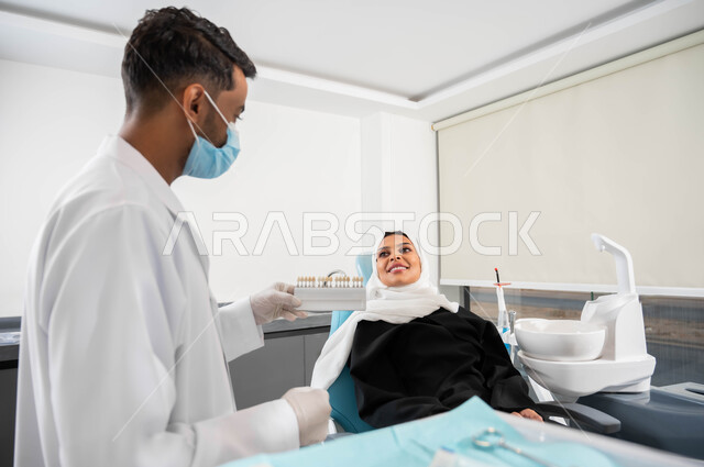 مفهوم الاهتمام والرعاية بصحة ومظهر الأسنان، طبيب أسنان يرتدي القفازات والكمامة الطبية يمسك بيده دليل الألوان لترميم أسنان المريضة، سيدة عربية خليجية سعودية محجبة في عيادة سنية تتلقى العلاج اللازم لصنع ابتسامة مميزة، مراجعة العيادات الصحية بشكل دوري